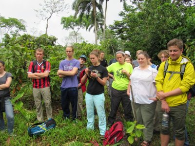 A group of about 15 students stands in a green forest listening to an unidentified speaker