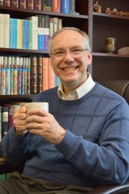 Ryan Ahlgrim wearing a blue sweater and holding a mug while sitting in front of a book shelf
