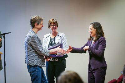 Two women shake hands while a third looks on.