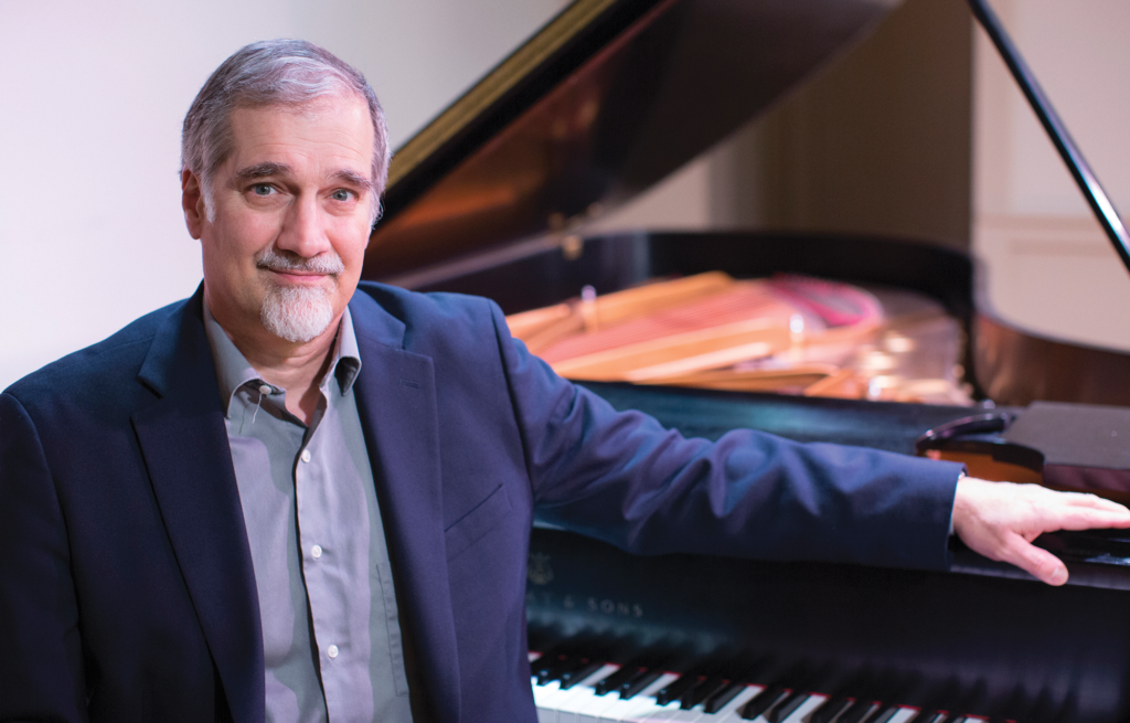 Man sitting in a blue suit with his arm resting on a piano ledge