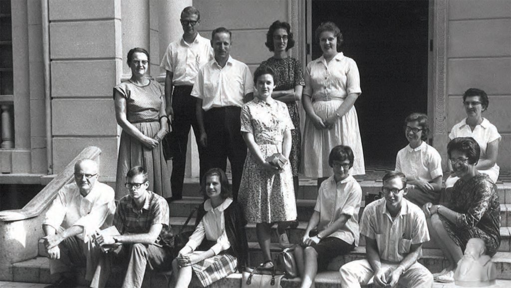 Black and white photo of fourteen people standing or sitting on steps