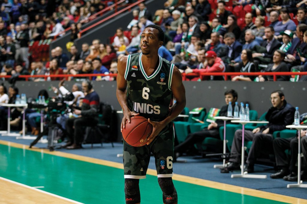 Man wearing a green basketball jersey holding a basketball. He is in a gymnasium with many people in the stands