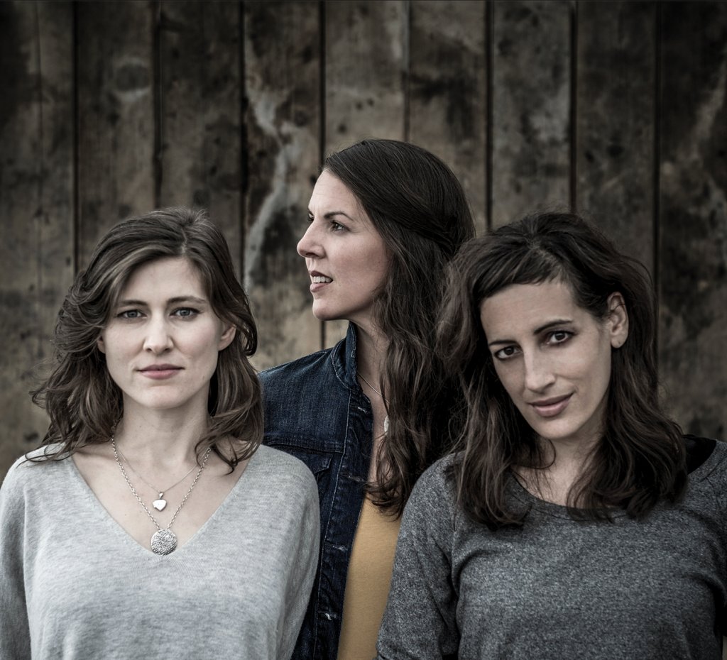 Three women. Two are facing forward and the third is looking off to the side. They are all standing in front of wooden planks forming a wall