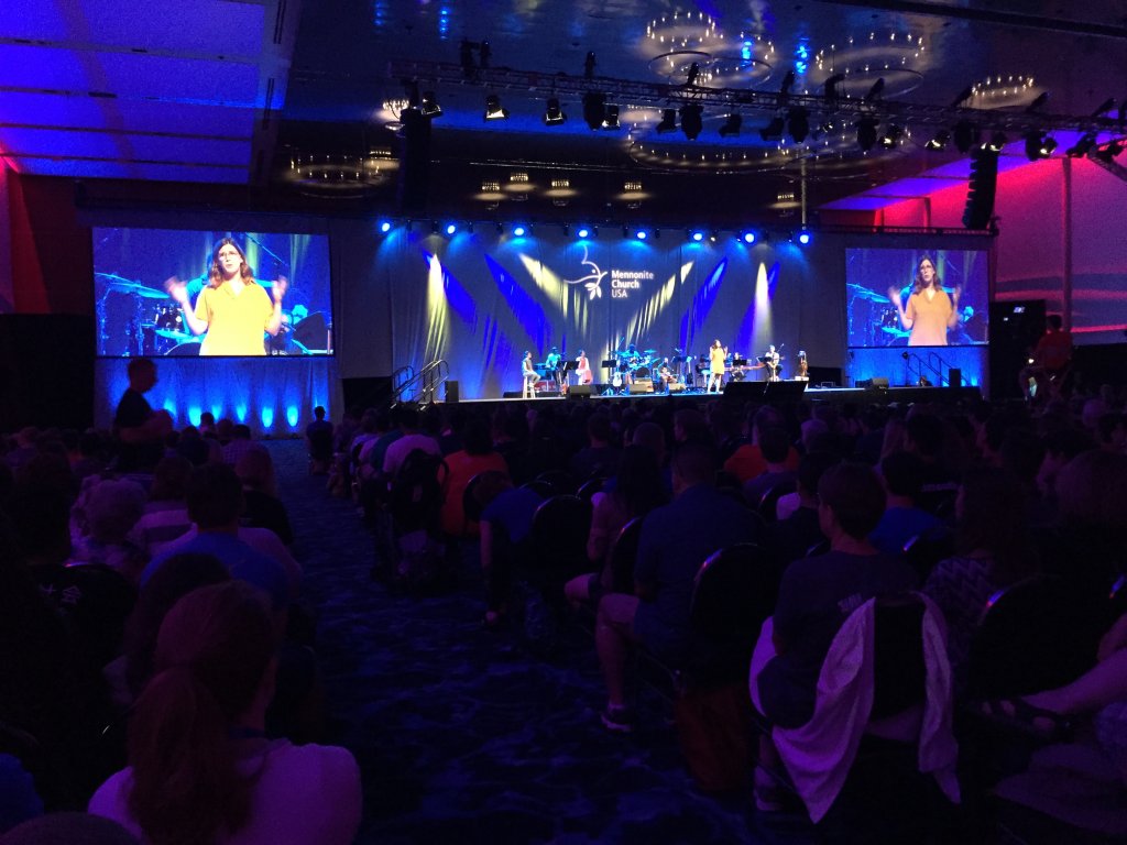 A large room full of people with a woman standing on a stage at the front of the room