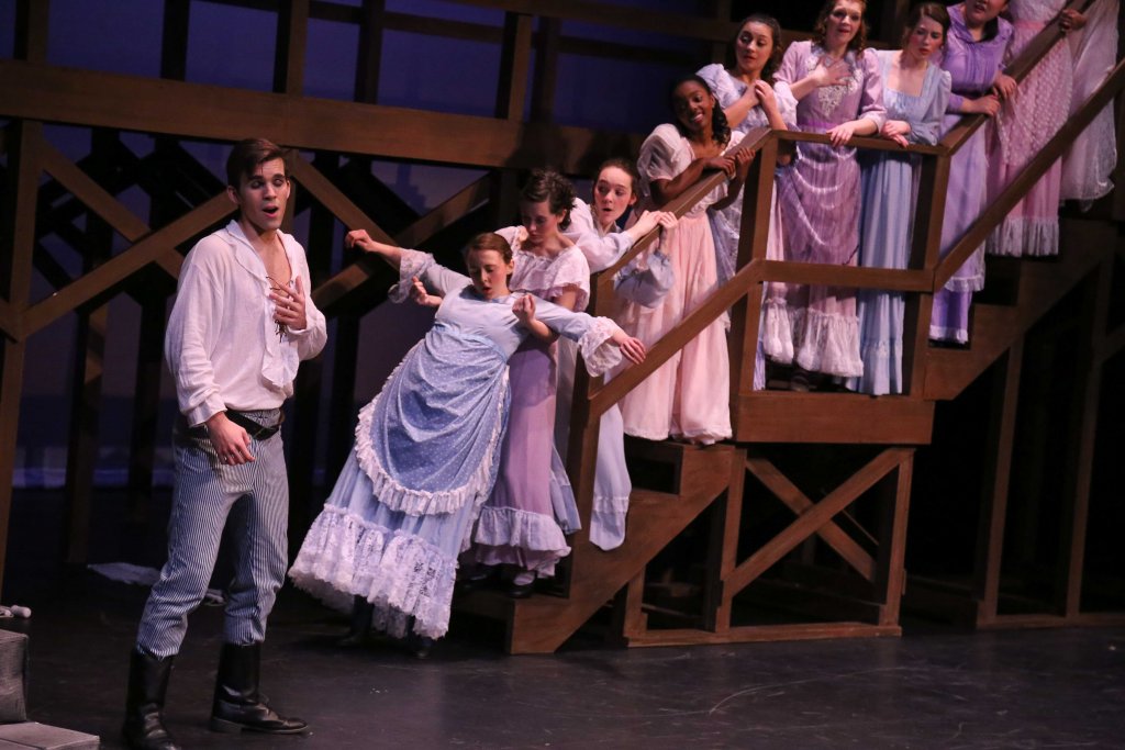 A man standing on stage with several woman standing on a set of stairs behind him