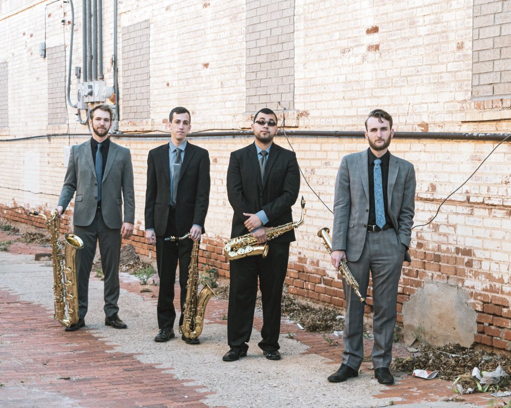 Four men in suits standing in front of a brick wall and holding brass instruments