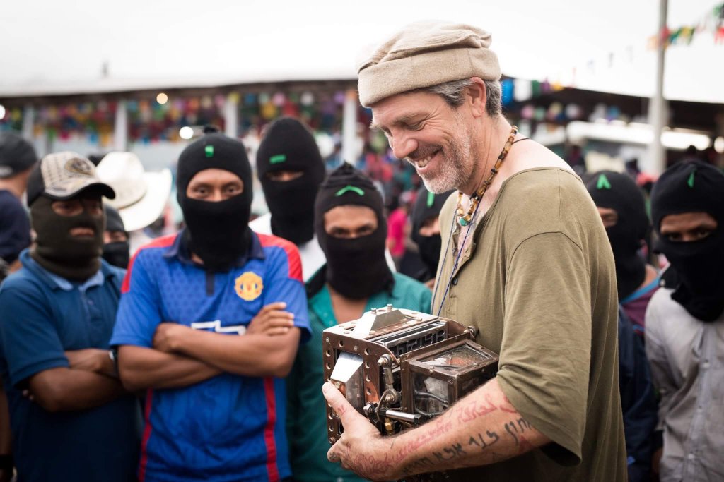 Man holding a contraption with men listening to him with covered faces