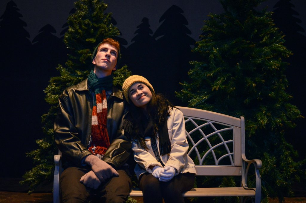 A man and a woman sitting on a bench in front of pine trees