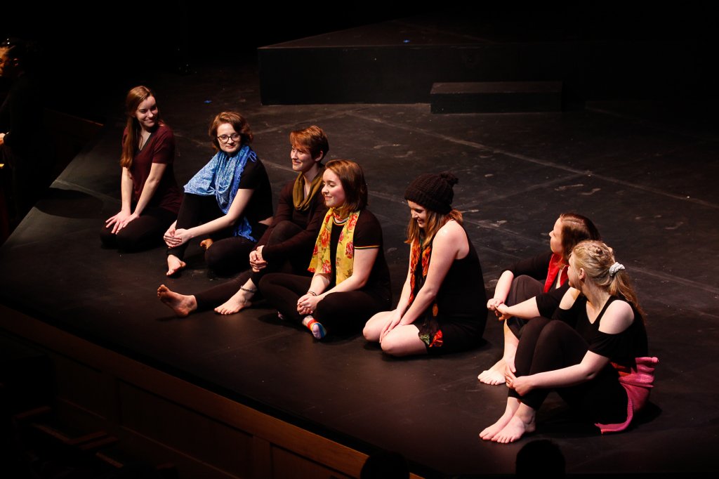 A group of woman barefoot wearing black and sitting in a line on stage